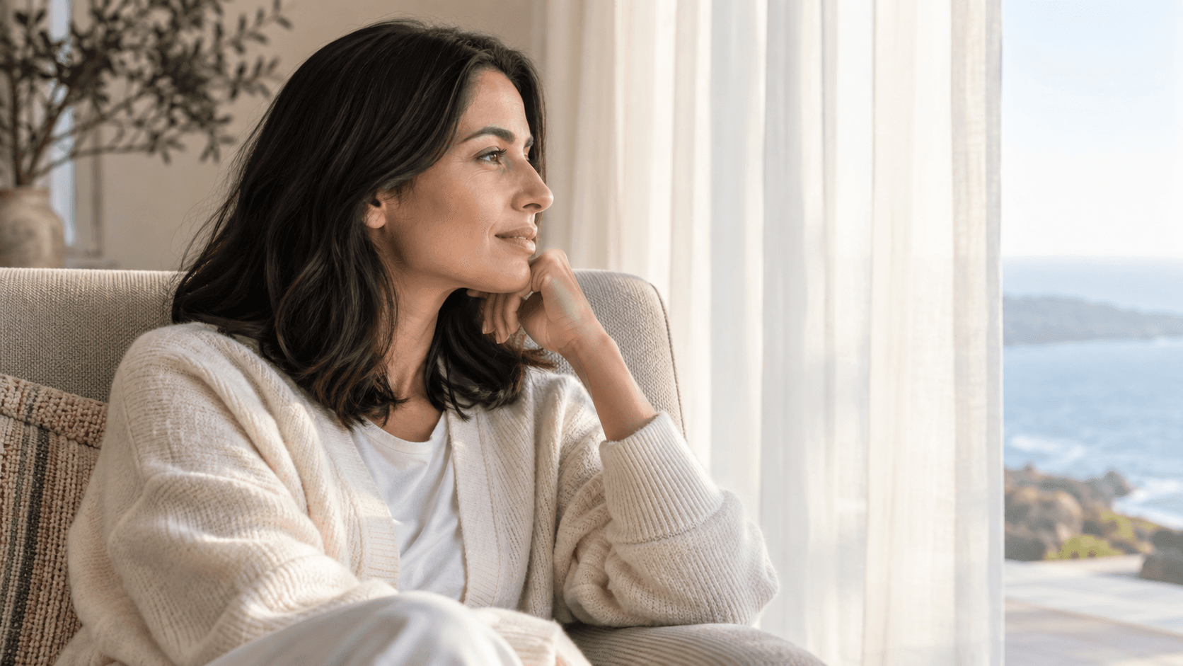 Woman in an ivory cardigan resting in an armchair beside a softly lit window