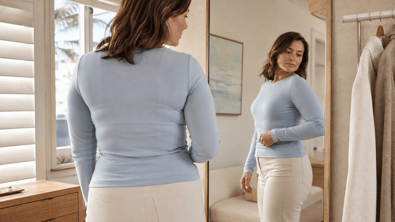 Fully clothed woman examining a natural bra-line contour beneath a fitted top in a private coastal room