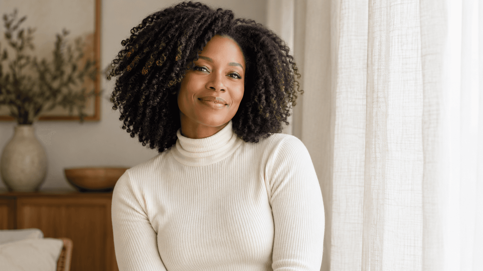 Woman with natural textured hair and a cream top beside a linen curtain.