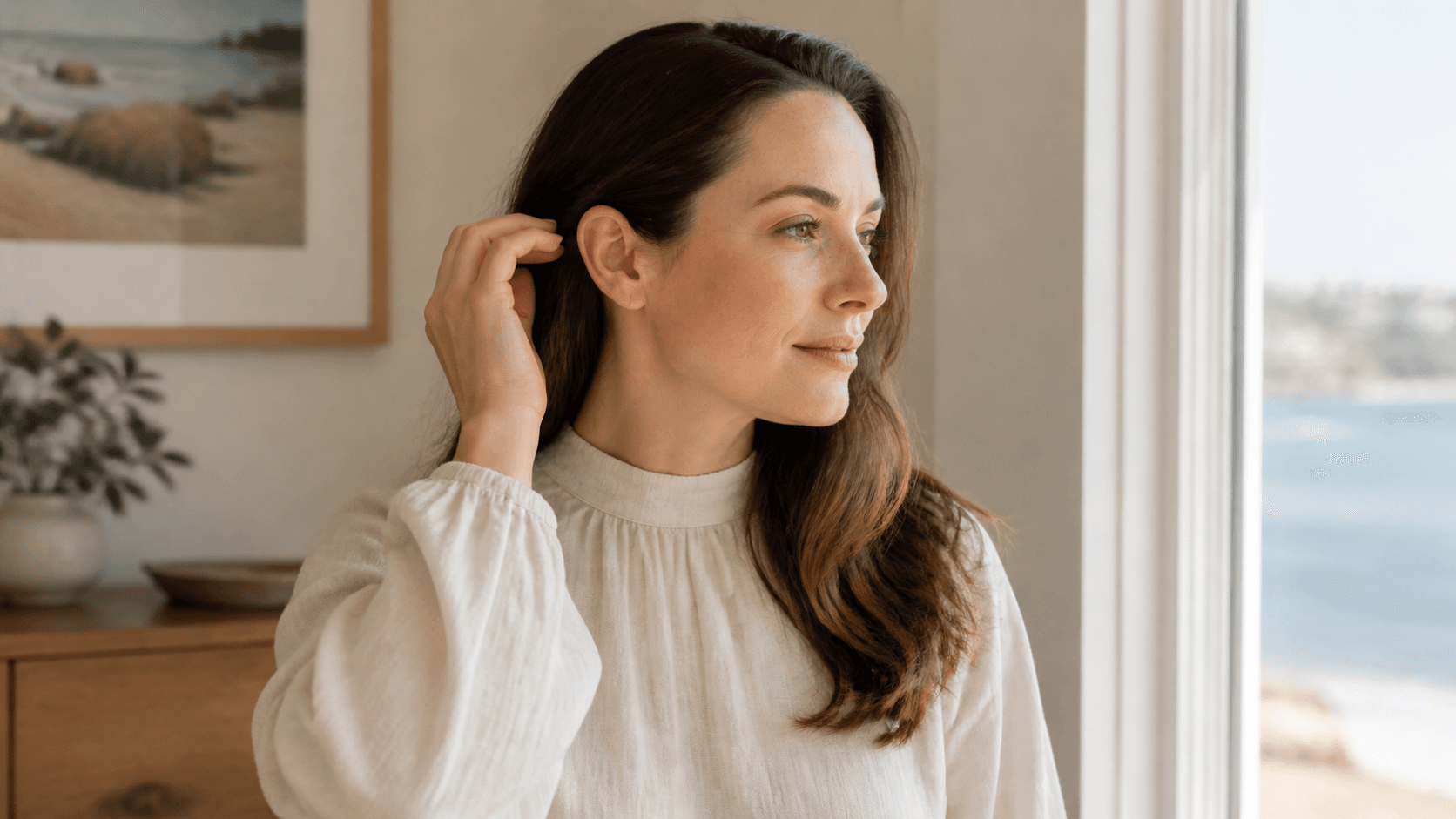Adult woman gently tucking her hair behind one ear in soft coastal light